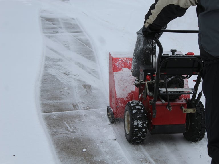 A person operating a snow blower to clear a concrete driveway covered in snow after a light winter storm, snow removal service in progress